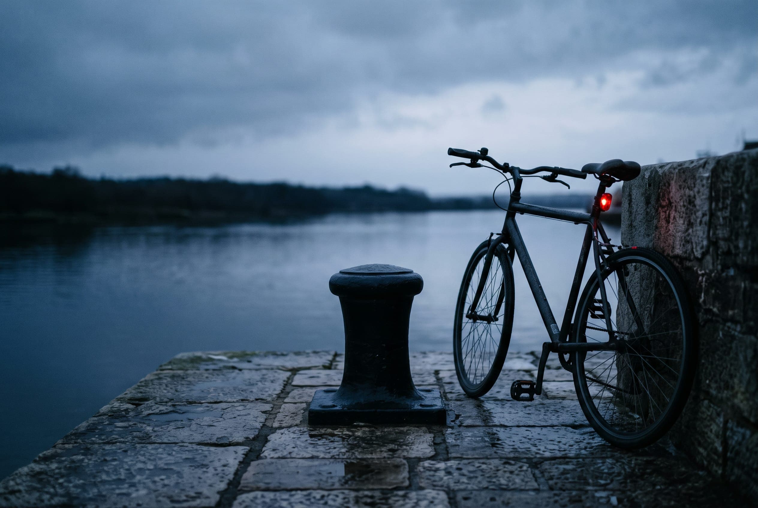 An iron mooring bollard on a wet quay at dusk, dark water just beyond the edge and a bicycle reflector catching the last light.