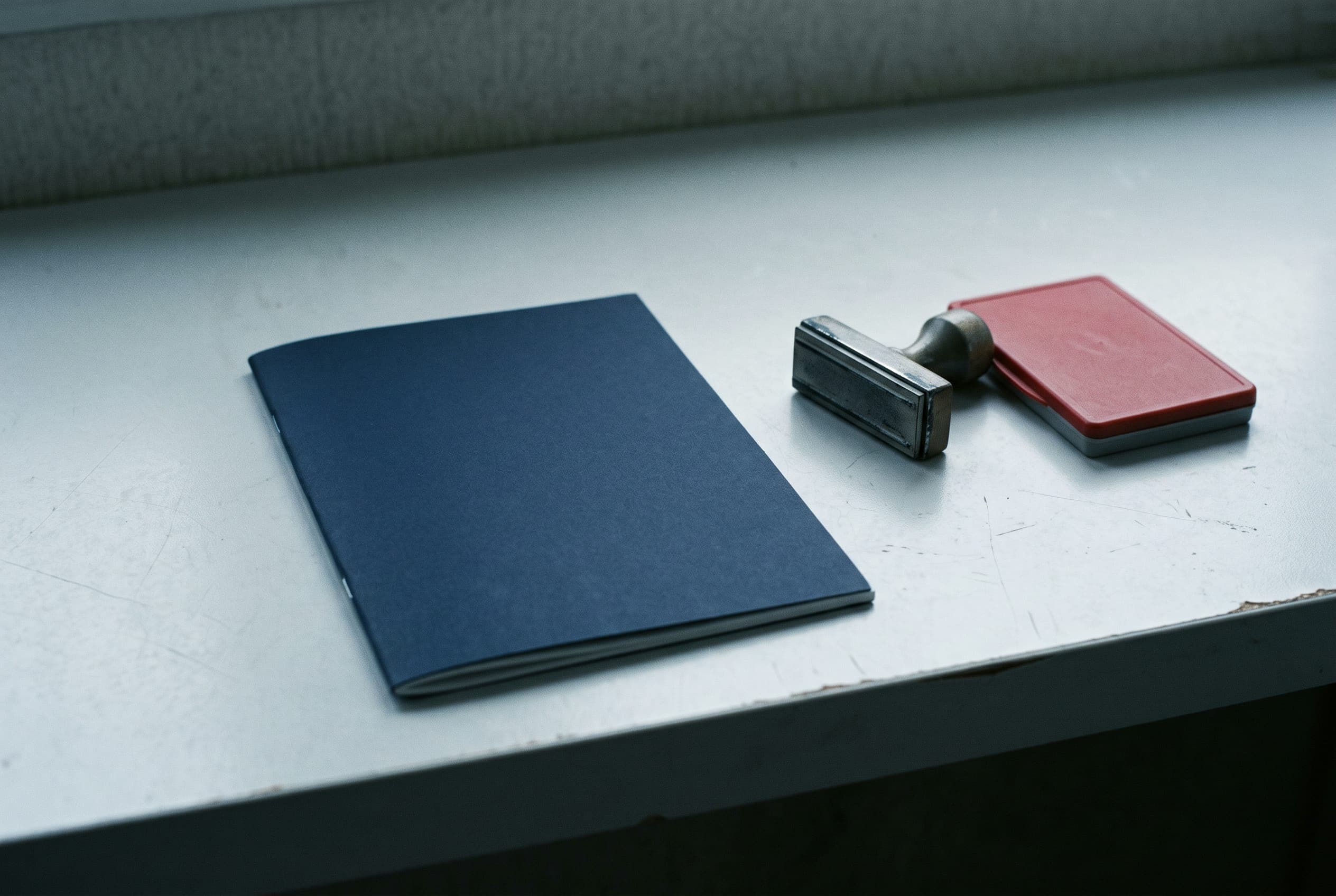 A plain navy booklet lying closed on a pale counter beside a steel desk stamp and a red inkpad.