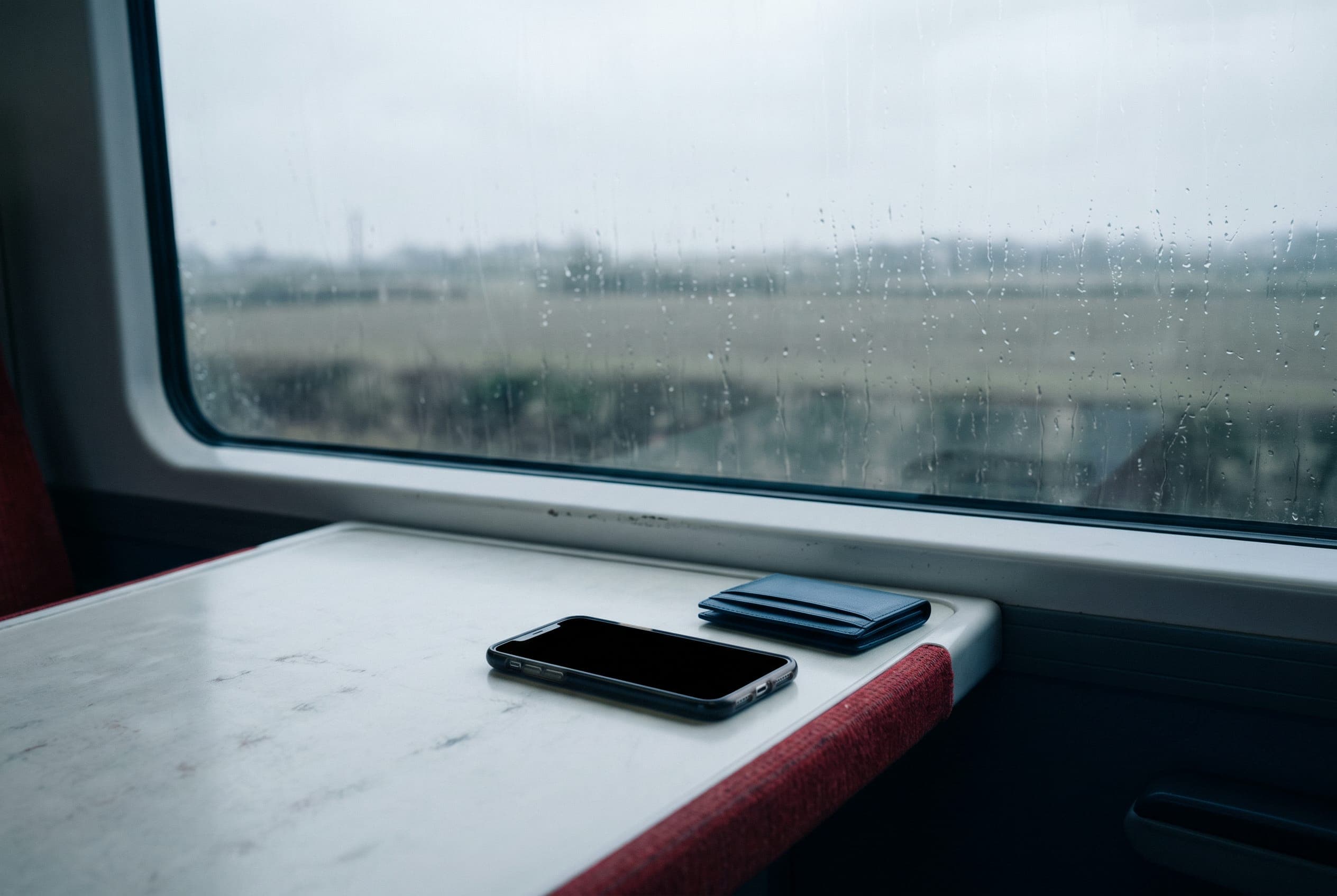 A phone with a blank screen and a slim card wallet on a train table, rain running down the window beside them.