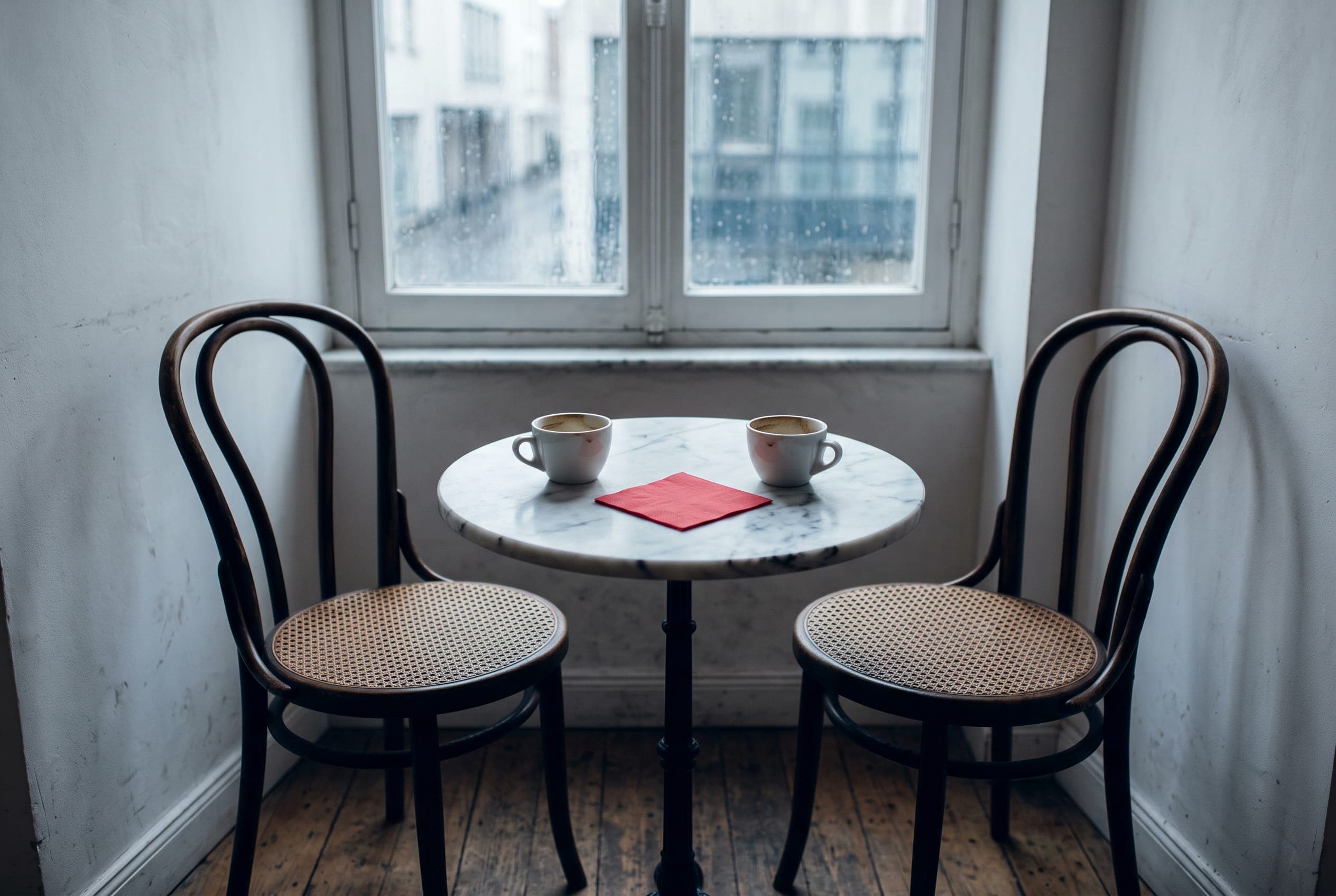 Two bentwood chairs facing each other across a small marble cafe table, two finished cups and a red napkin between them.