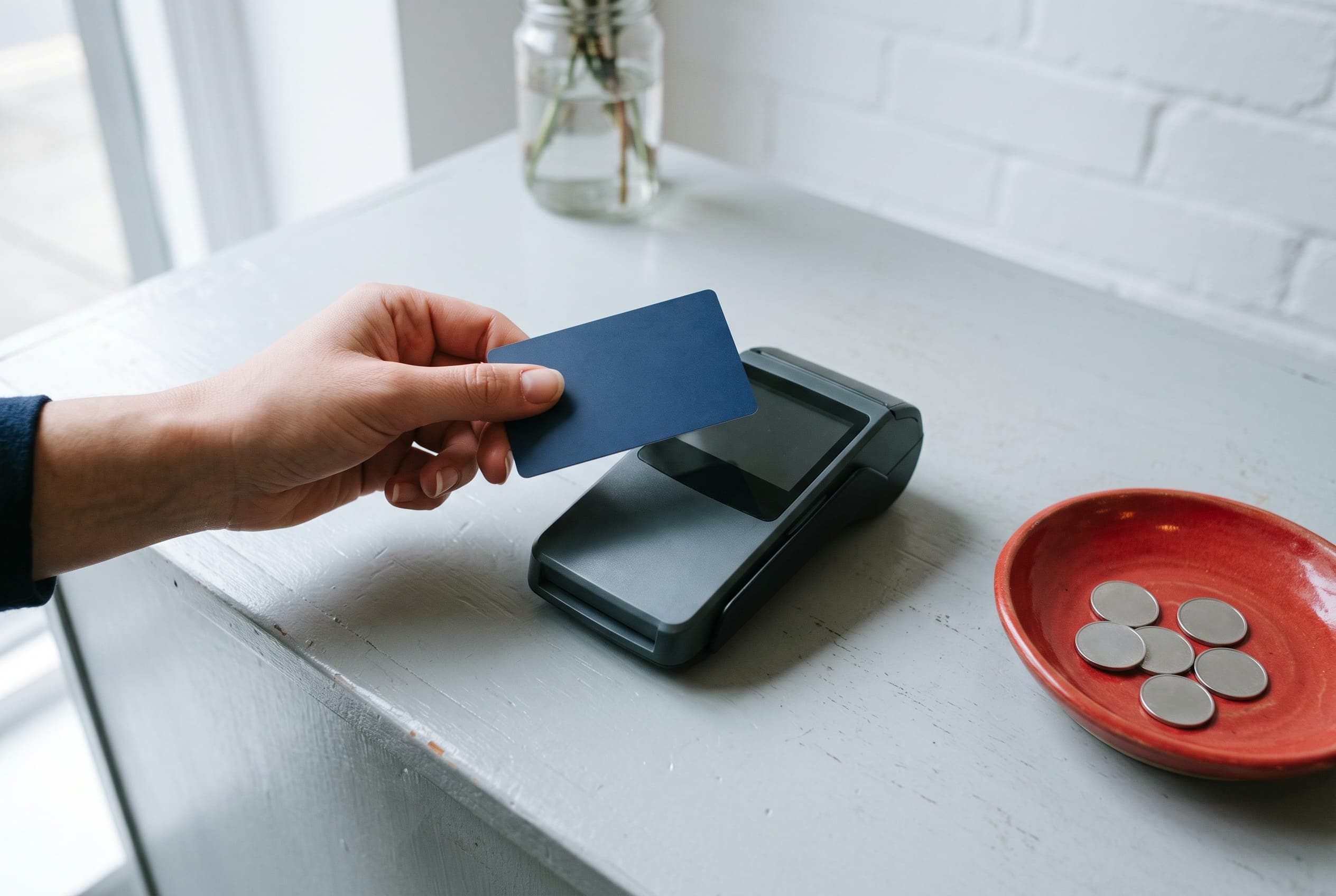 A hand holding a plain navy card above a payment terminal with a dark screen, a red dish of coins beside it.